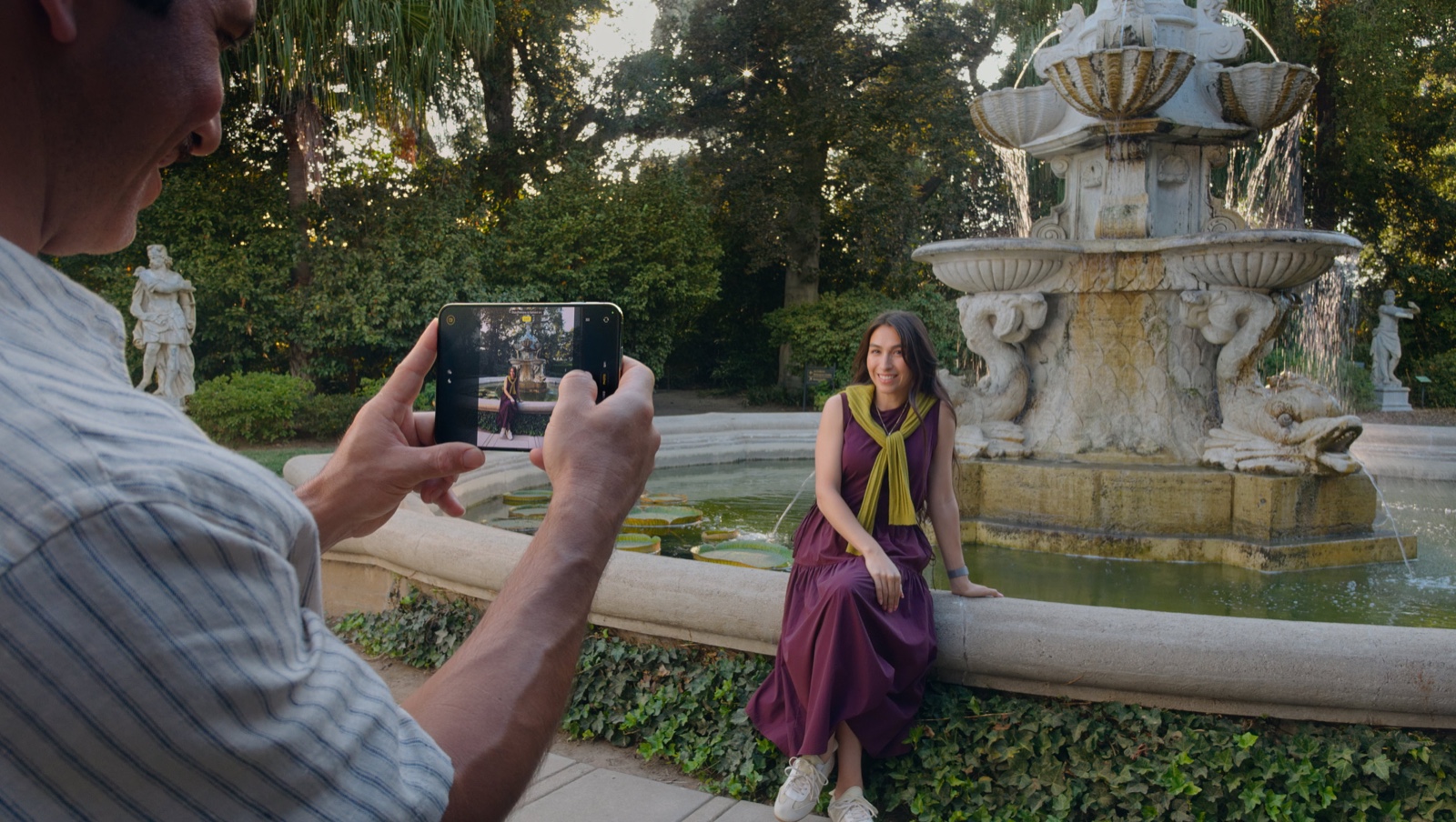A man photographing a woman sitting on a fountain edge with an iPhone Duo, seeing the live frame on its screen. Photo: Apple